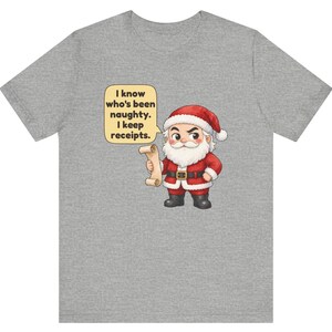 May include: A heather gray t-shirt featuring a cartoon Santa Claus holding a scroll. A yellow speech bubble next to him contains the black text: I know who's been naughty. I keep receipts. The Santa wears a red suit with white trim and black boots.
