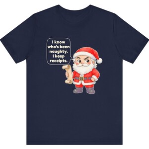 May include: A navy blue t-shirt featuring a cartoon illustration of Santa Claus holding a scroll. A speech bubble next to Santa contains the text: I know who's been naughty. I keep receipts. Santa wears a red suit with white trim and a black belt.