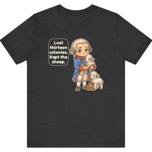 Peut inclure: Un t-shirt gris anthracite chin&eacute; avec une illustration de style dessin anim&eacute; repr&eacute;sentant un personnage historique en manteau bleu tenant un agneau, accompagn&eacute; d'un autre agneau et d'une petite horloge. Une bulle contient le texte : Lost thirteen colonies. Kept the sheep.