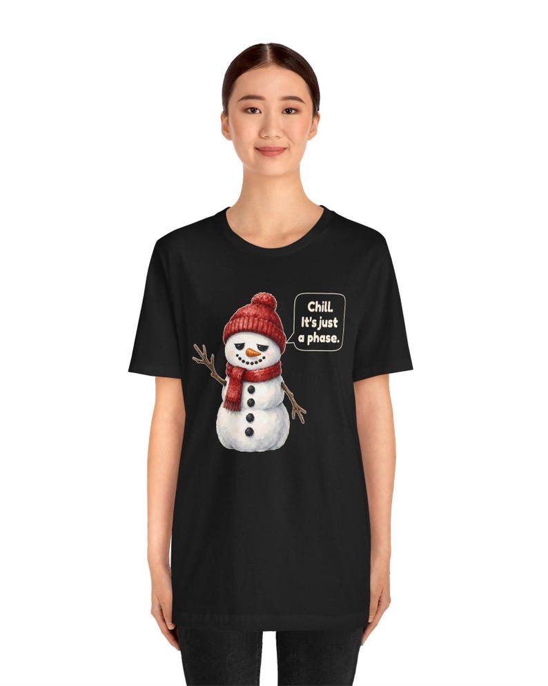 Pode incluir: Uma t-shirt preta com um gr&aacute;fico de um boneco de neve a usar um gorro e um cachecol de malha vermelhos. O boneco de neve tem um nariz de cenoura, bra&ccedil;os de ramo e um bal&atilde;o de fala com o texto: Chill. It's just a phase.