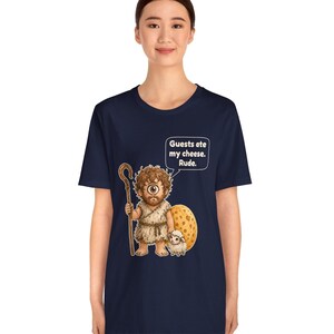 May include: A navy blue t-shirt featuring a graphic of a cyclops holding a wooden staff and standing next to a small sheep and a large wheel of cheese. A speech bubble above the cyclops contains the text: Guests ate my cheese. Rude.
