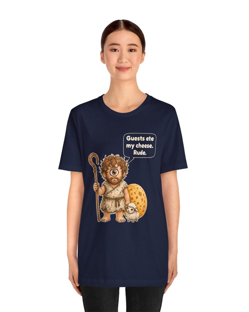 May include: A navy blue t-shirt featuring a graphic of a cyclops holding a wooden staff and standing next to a small sheep and a large wheel of cheese. A speech bubble above the cyclops contains the text: Guests ate my cheese. Rude.