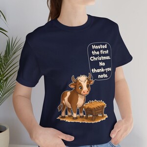 以下が含まれることがあります： ネイビーブルーのTシャツ。干し草が入った木製の飼い葉桶の横に立つ、茶色と白の子牛の漫画イラストが描かれています。子牛の上にある吹き出しには、白い文字で「Hosted the first Christmas. No thank-you note.」と書かれています。