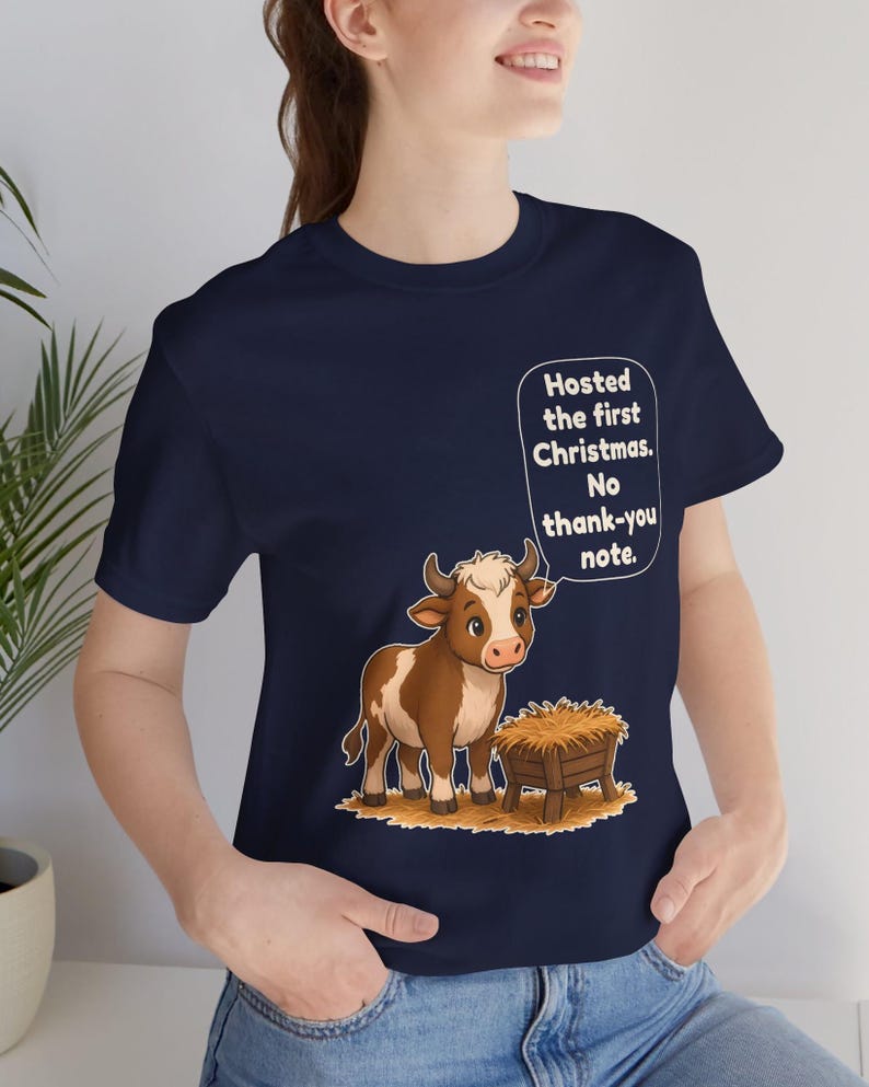 以下が含まれることがあります： ネイビーブルーのTシャツ。干し草が入った木製の飼い葉桶の横に立つ、茶色と白の子牛の漫画イラストが描かれています。子牛の上にある吹き出しには、白い文字で「Hosted the first Christmas. No thank-you note.」と書かれています。