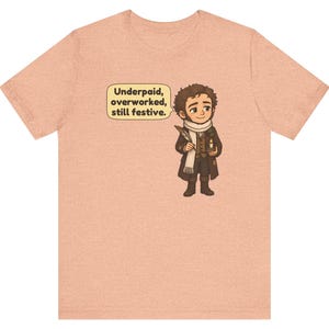 May include: A peach-colored short-sleeve t-shirt featuring a cartoon illustration of a person in Victorian-style clothing holding a quill and candle. A speech bubble contains the text: Underpaid, overworked, still festive.
