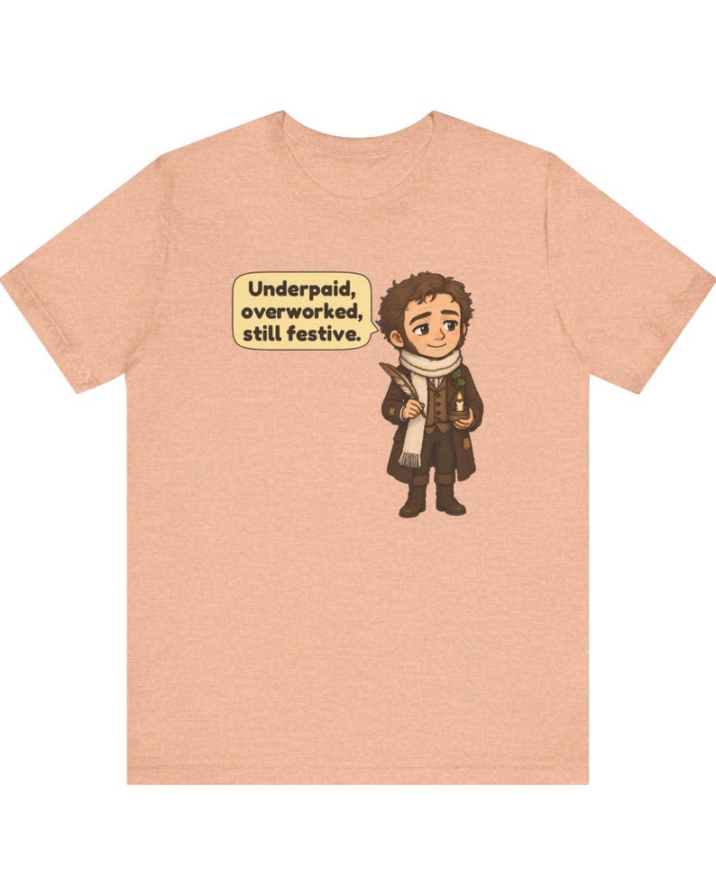 May include: A peach-colored short-sleeve t-shirt featuring a cartoon illustration of a person in Victorian-style clothing holding a quill and candle. A speech bubble contains the text: Underpaid, overworked, still festive.