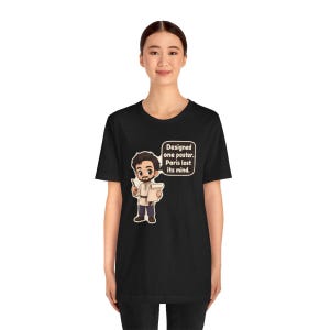 May include: A black short-sleeve t-shirt featuring a cartoon illustration of a person holding a pen and paper with a speech bubble that reads: Designed one poster. Paris lost its mind. The graphic is centered on the chest of the shirt.