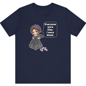 May include: A navy blue t-shirt featuring a graphic of a character in a black dress with a lilac floral headpiece and a fan. A speech bubble contains the text: Everyone wore lilac. I wore black. A small lilac ribbon is positioned below.