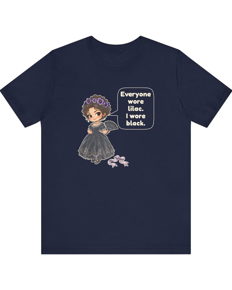 May include: A navy blue t-shirt featuring a graphic of a character in a black dress with a lilac floral headpiece and a fan. A speech bubble contains the text: Everyone wore lilac. I wore black. A small lilac ribbon is positioned below.