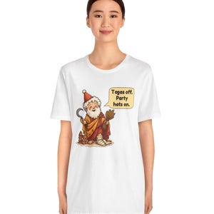 May include: A white t-shirt featuring a cartoon illustration of a bearded figure wearing a red party hat and a toga, holding a sickle and a bundle of wheat. A speech bubble next to the figure contains the text: Togas off. Party hats on.