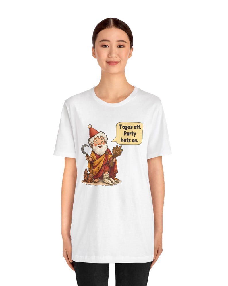 May include: A white t-shirt featuring a cartoon illustration of a bearded figure wearing a red party hat and a toga, holding a sickle and a bundle of wheat. A speech bubble next to the figure contains the text: Togas off. Party hats on.