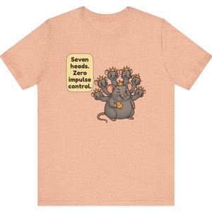 May include: A peach-colored t-shirt featuring a graphic of a gray seven-headed rat wearing small gold crowns and holding a piece of yellow cheese. A speech bubble next to the rat contains the text: Seven heads. Zero impulse control.