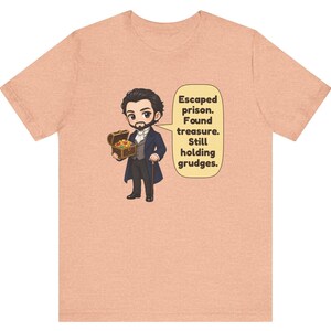 May include: A peach-colored t-shirt featuring a cartoon illustration of a man in a dark coat holding a treasure chest. A speech bubble next to the character contains the text: Escaped prison. Found treasure. Still holding grudges.