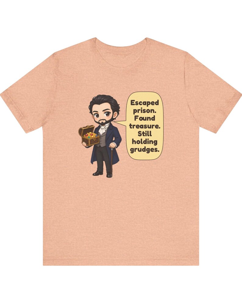 May include: A peach-colored t-shirt featuring a cartoon illustration of a man in a dark coat holding a treasure chest. A speech bubble next to the character contains the text: Escaped prison. Found treasure. Still holding grudges.