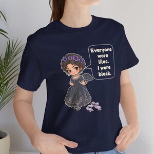 May include: A navy blue t-shirt featuring a graphic of a character in a black dress with a purple flower crown and a hand fan. A speech bubble contains the text: Everyone wore lilac. I wore black. A small purple ribbon is below the character.