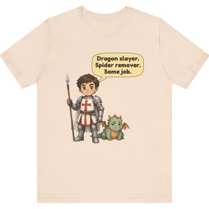 K&ouml;nnte beinhalten: Ein hellbeiges T-Shirt mit einer Cartoon-Illustration eines Ritters in silberner R&uuml;stung, der einen Speer h&auml;lt und neben einem kleinen gr&uuml;nen Drachen steht. Eine Sprechblase &uuml;ber dem Ritter enth&auml;lt den Text: Dragon slayer. Spider remover. Same job.