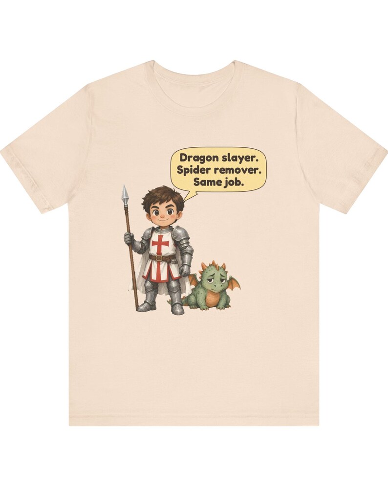 K&ouml;nnte beinhalten: Ein hellbeiges T-Shirt mit einer Cartoon-Illustration eines Ritters in silberner R&uuml;stung, der einen Speer h&auml;lt und neben einem kleinen gr&uuml;nen Drachen steht. Eine Sprechblase &uuml;ber dem Ritter enth&auml;lt den Text: Dragon slayer. Spider remover. Same job.