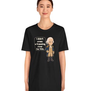 May include: A black t-shirt featuring a cartoon illustration of a historical figure in a blue and tan uniform. A speech bubble next to the character contains the white text: I didn't cross a freezing river for this.