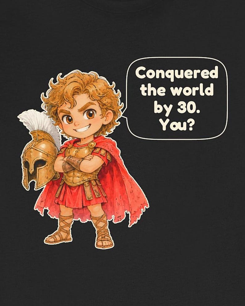 May include: A black t-shirt featuring a cartoon illustration of a curly-haired figure in bronze armor and a red cape, holding a plumed helmet. A speech bubble contains the text: Conquered the world by 30. You?