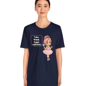 May include: A navy blue t-shirt featuring a graphic of a cartoon ballerina with pink hair, a pink tutu, and a star wand. A speech bubble next to the character contains the text: Tutu. Wand. Sugar addiction. The design is centered on the chest.