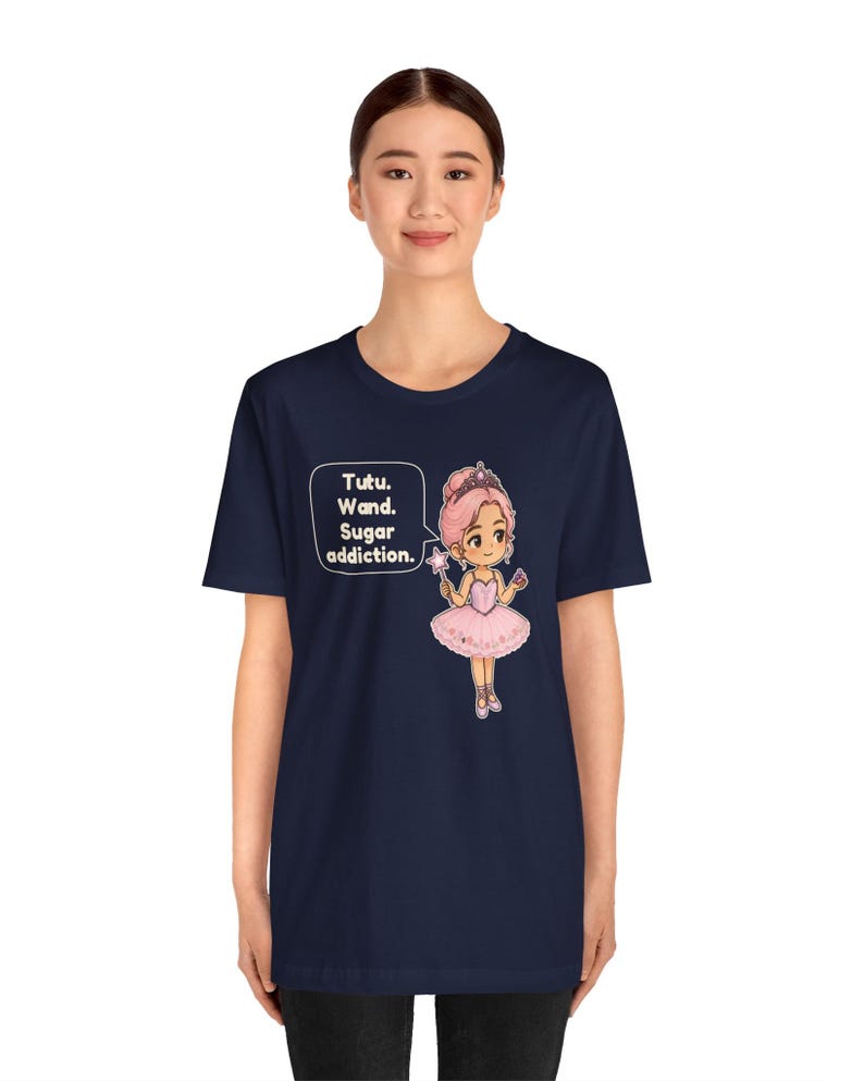 May include: A navy blue t-shirt featuring a graphic of a cartoon ballerina with pink hair, a pink tutu, and a star wand. A speech bubble next to the character contains the text: Tutu. Wand. Sugar addiction. The design is centered on the chest.
