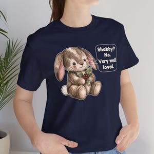 May include: A navy blue t-shirt featuring a graphic of a brown, stitched plush rabbit holding a sprig of holly with red berries. A speech bubble next to the rabbit contains the text: Shabby? No. Very well loved.