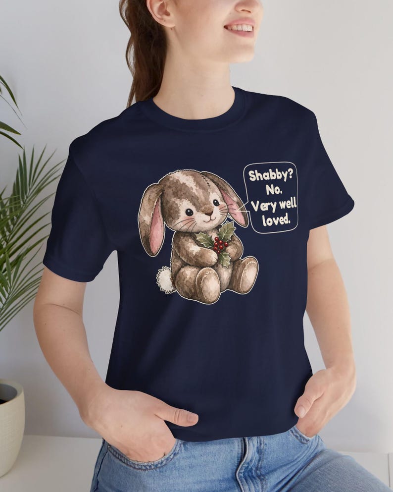 May include: A navy blue t-shirt featuring a graphic of a brown, stitched plush rabbit holding a sprig of holly with red berries. A speech bubble next to the rabbit contains the text: Shabby? No. Very well loved.