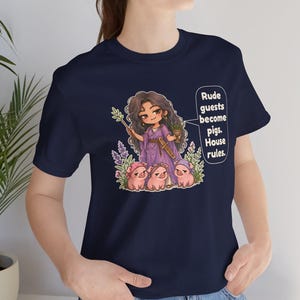 May include: A navy blue t-shirt featuring a graphic illustration of a long-haired character in a purple robe holding a wand and a goblet, surrounded by three small pigs. The text reads: Rude guests become pigs. House rules.