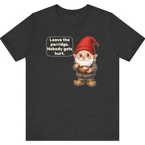 May include: A dark charcoal grey t-shirt featuring a graphic of a gnome wearing a red hat and brown clothing, holding a wooden bowl and spoon. A speech bubble next to the gnome contains the text: Leave the porridge. Nobody gets hurt.