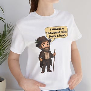 May include: A white short-sleeved t-shirt featuring a cartoon illustration of a bearded traveler wearing a brown hat with a pine sprig, a brown coat, and boots, holding a wooden walking stick. A speech bubble contains the text: I walked a thousand miles. Pack a lunch.