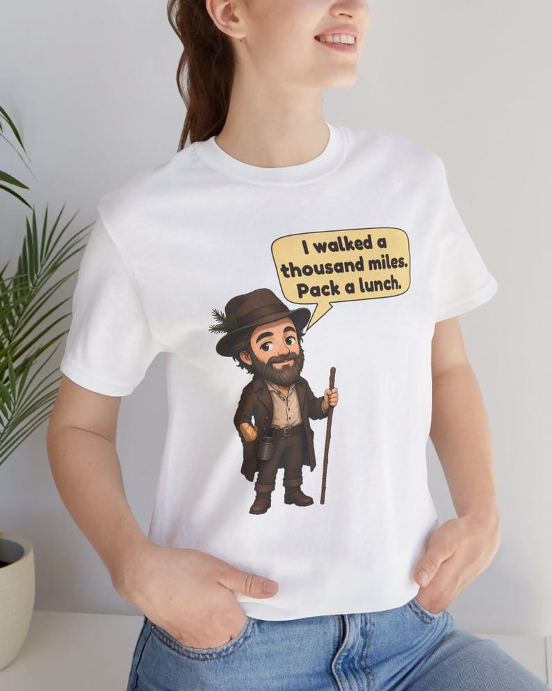 May include: A white short-sleeved t-shirt featuring a cartoon illustration of a bearded traveler wearing a brown hat with a pine sprig, a brown coat, and boots, holding a wooden walking stick. A speech bubble contains the text: I walked a thousand miles. Pack a lunch.