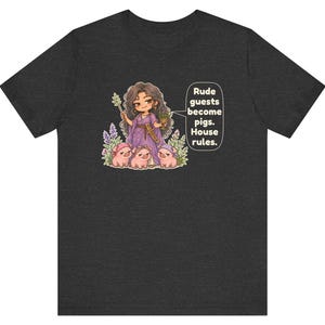 May include: A dark heather gray t-shirt featuring a cartoon illustration of a character in a purple dress holding a staff and cup, surrounded by three small pink pigs. A speech bubble contains the text: Rude guests become pigs. House rules.