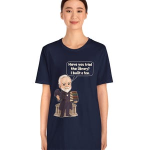 May include: A navy blue short-sleeve t-shirt featuring a cartoon illustration of a bearded man in a suit holding a stack of books in front of a building. A speech bubble contains the text: Have you tried the library? I built a few.