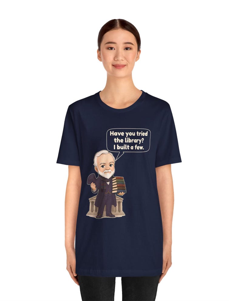 May include: A navy blue short-sleeve t-shirt featuring a cartoon illustration of a bearded man in a suit holding a stack of books in front of a building. A speech bubble contains the text: Have you tried the library? I built a few.