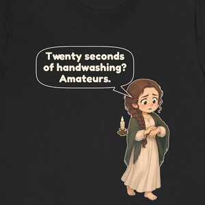 May include: A black t-shirt featuring a cartoon illustration of a person with long brown braided hair wearing a cream dress and dark shawl. The person holds a lit candle and a speech bubble reads: Twenty seconds of handwashing? Amateurs.