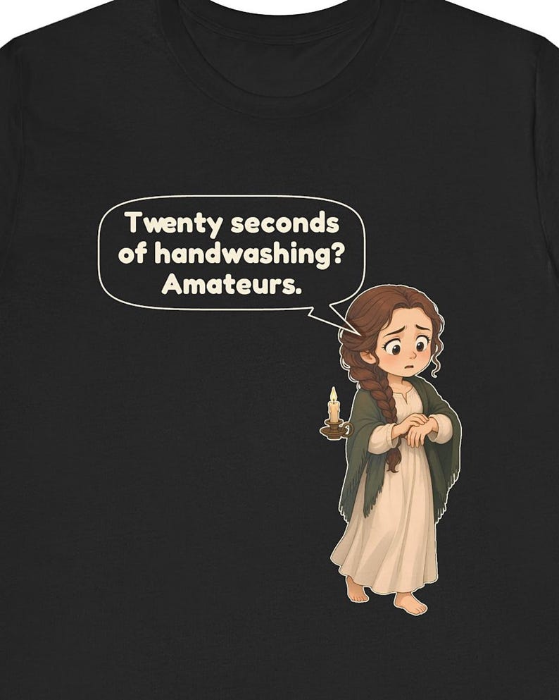 Op de afbeelding: Een zwart t-shirt met een cartoonillustratie van een persoon met lang bruin gevlochten haar in een cr&egrave;mekleurige jurk en een donkere sjaal. De persoon houdt een brandende kaars vast en een tekstballon zegt: Twenty seconds of handwashing? Amateurs.