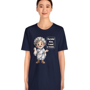 May include: A navy blue t-shirt featuring a cartoon illustration of a bearded figure with curly gray hair wearing a white toga and brown sandals. The figure holds a white cloth and has a speech bubble that reads: Eureka! Also, I need a towel.