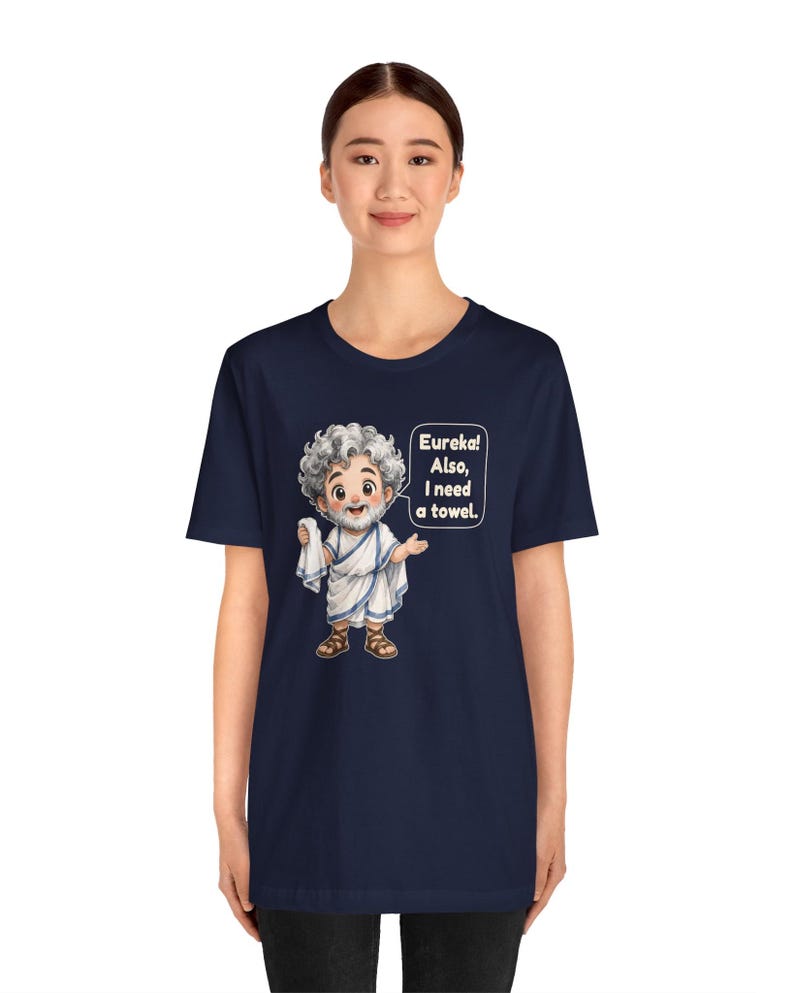 May include: A navy blue t-shirt featuring a cartoon illustration of a bearded figure with curly gray hair wearing a white toga and brown sandals. The figure holds a white cloth and has a speech bubble that reads: Eureka! Also, I need a towel.