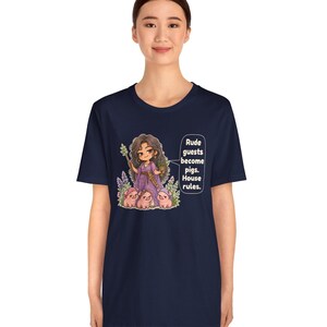 May include: A navy blue t-shirt featuring a graphic of a character with long dark hair in a purple dress holding a staff and a potted plant, surrounded by three small pigs. The text reads: Rude guests become pigs. House rules.