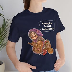Puede incluir: Una camiseta azul marino con el gr&aacute;fico de una persona mayor con pa&ntilde;uelo y gafas montada en una escoba. La persona lleva un saco y un calcet&iacute;n rojo. Un bocadillo de texto contiene: Sweeping in late. Fashionably.