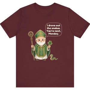 Puede incluir: Una camiseta de manga corta color granate con una ilustraci&oacute;n de dibujos animados de un obispo sosteniendo un bast&oacute;n de madera y un tr&eacute;bol. Una peque&ntilde;a serpiente verde est&aacute; cerca. Un globo de texto contiene: I drove out the snakes. You're next, Monday.