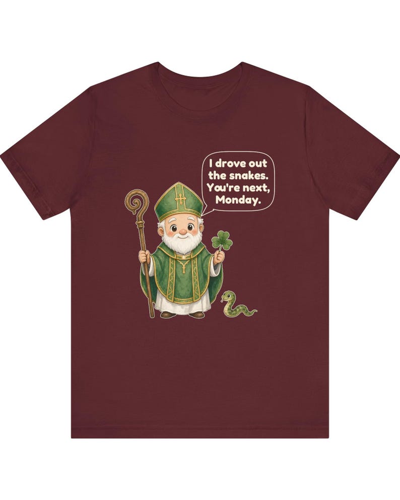 Puede incluir: Una camiseta de manga corta color granate con una ilustraci&oacute;n de dibujos animados de un obispo sosteniendo un bast&oacute;n de madera y un tr&eacute;bol. Una peque&ntilde;a serpiente verde est&aacute; cerca. Un globo de texto contiene: I drove out the snakes. You're next, Monday.