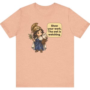 Puede incluir: Una camiseta color melocot&oacute;n con el gr&aacute;fico de un personaje con casco dorado y t&uacute;nica azul que sostiene una rama de olivo y un libro con un b&uacute;ho. Un bocadillo contiene el texto: Show your work. The owl is watching.