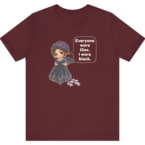 May include: A maroon short-sleeve t-shirt featuring a cartoon illustration of a character in a black dress with a lilac flower crown and fan. A speech bubble contains the text: Everyone wore lilac. I wore black. A small lilac ribbon is below.