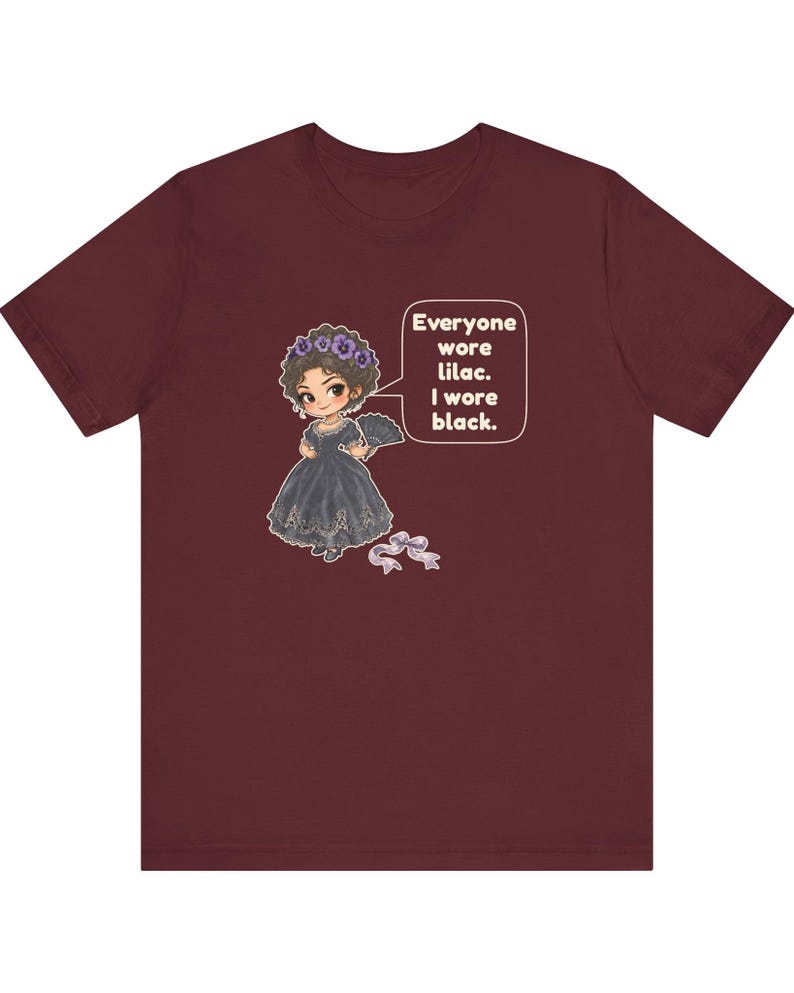 May include: A maroon short-sleeve t-shirt featuring a cartoon illustration of a character in a black dress with a lilac flower crown and fan. A speech bubble contains the text: Everyone wore lilac. I wore black. A small lilac ribbon is below.