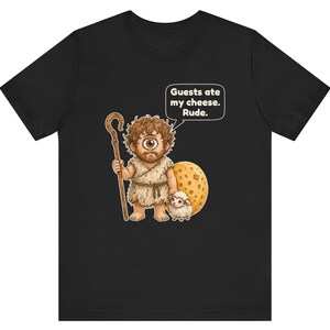 May include: A black t-shirt featuring a graphic of a one-eyed cyclops holding a wooden staff next to a small white sheep and a large wheel of Swiss cheese. A speech bubble above the cyclops contains the text: Guests ate my cheese. Rude.