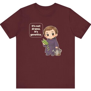 May include: A maroon t-shirt featuring a cartoon illustration of a monk holding a pea pod and a watering can. A speech bubble next to the character contains the text: It's not drama. It's genetics. The design is centered on the front of the shirt.