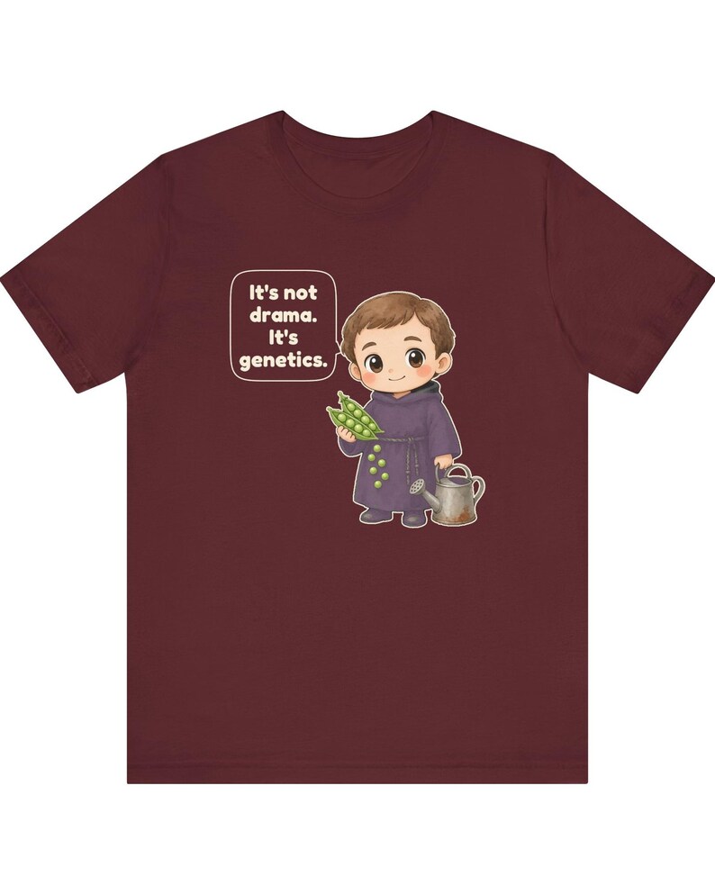 May include: A maroon t-shirt featuring a cartoon illustration of a monk holding a pea pod and a watering can. A speech bubble next to the character contains the text: It's not drama. It's genetics. The design is centered on the front of the shirt.