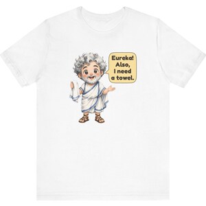 May include: A white short-sleeve t-shirt featuring a cartoon illustration of a bearded man in a white toga and brown sandals holding a white towel. A yellow speech bubble next to the character contains the text: Eureka! Also, I need a towel.