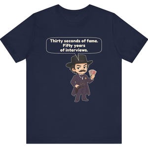 Pu&ograve; includere: Una t-shirt blu navy con un'illustrazione a cartoni animati di un personaggio con baffi, cappello e cappotto neri, che tiene in mano carte da gioco e una pistola. Un fumetto sopra il personaggio contiene il testo: Thirty seconds of fame. Fifty years of interviews.